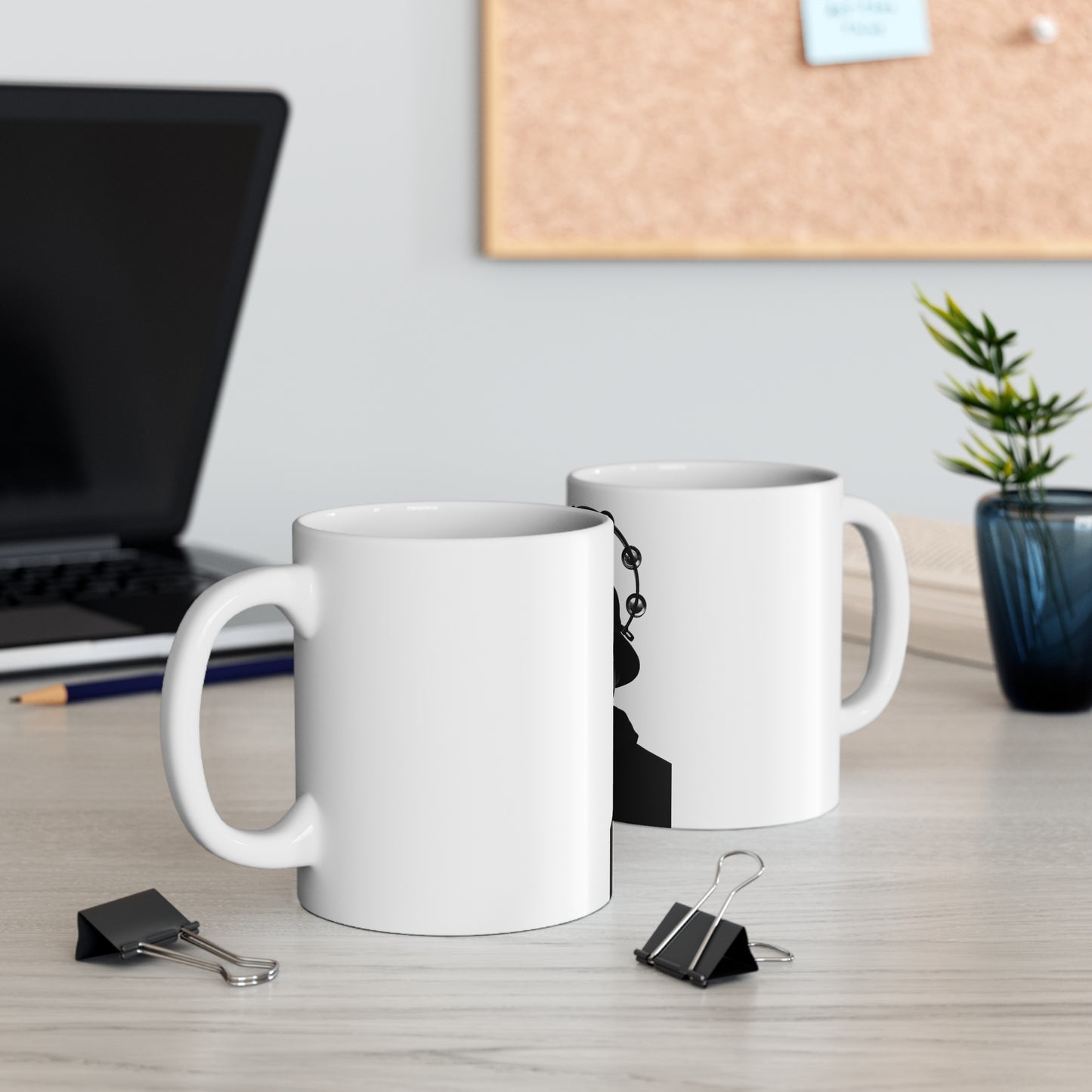 Two white coffee mugs on wooden office desk with laptop, pencil, paper clips, small plant, and corkboard