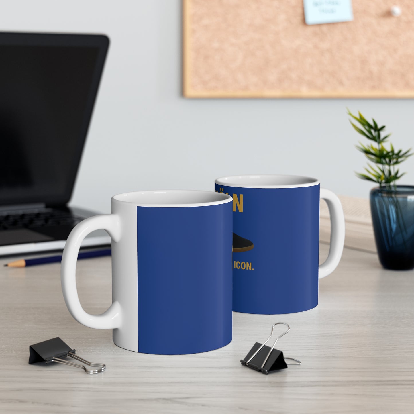 Two white ceramic mugs with blue outside, black binder clips on desk, laptop and plant in background