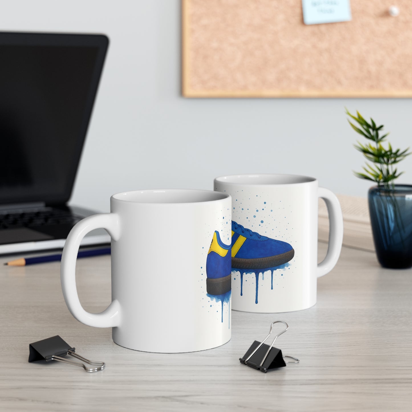White ceramic coffee mugs with blue and yellow sneaker design on a light wood desk with laptop and plant