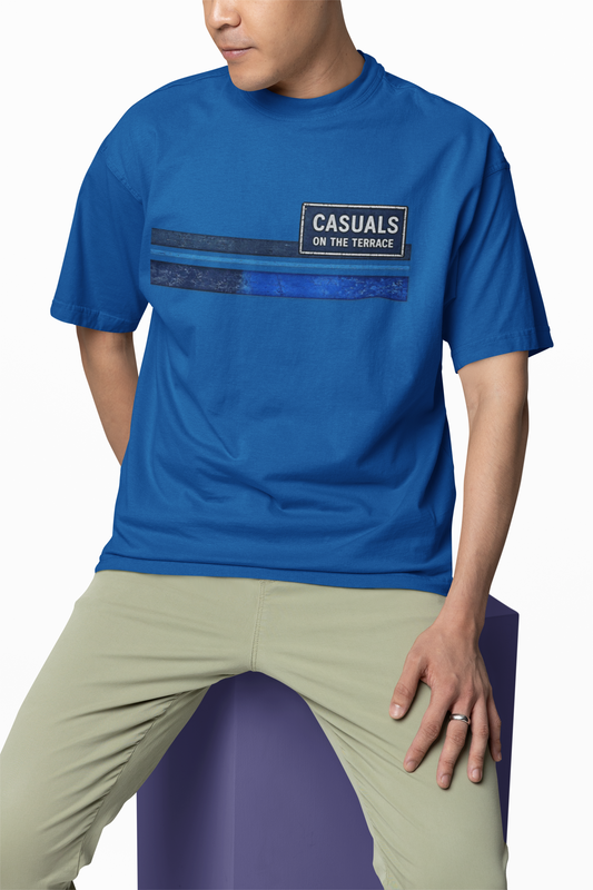 A man wearing a royal blue short-sleeve t-shirt with a graphic print on the chest that reads 'CASUALS ON THE TERRACE' over dark blue stripes, paired with light olive green pants, seated on a purple block against a white background.