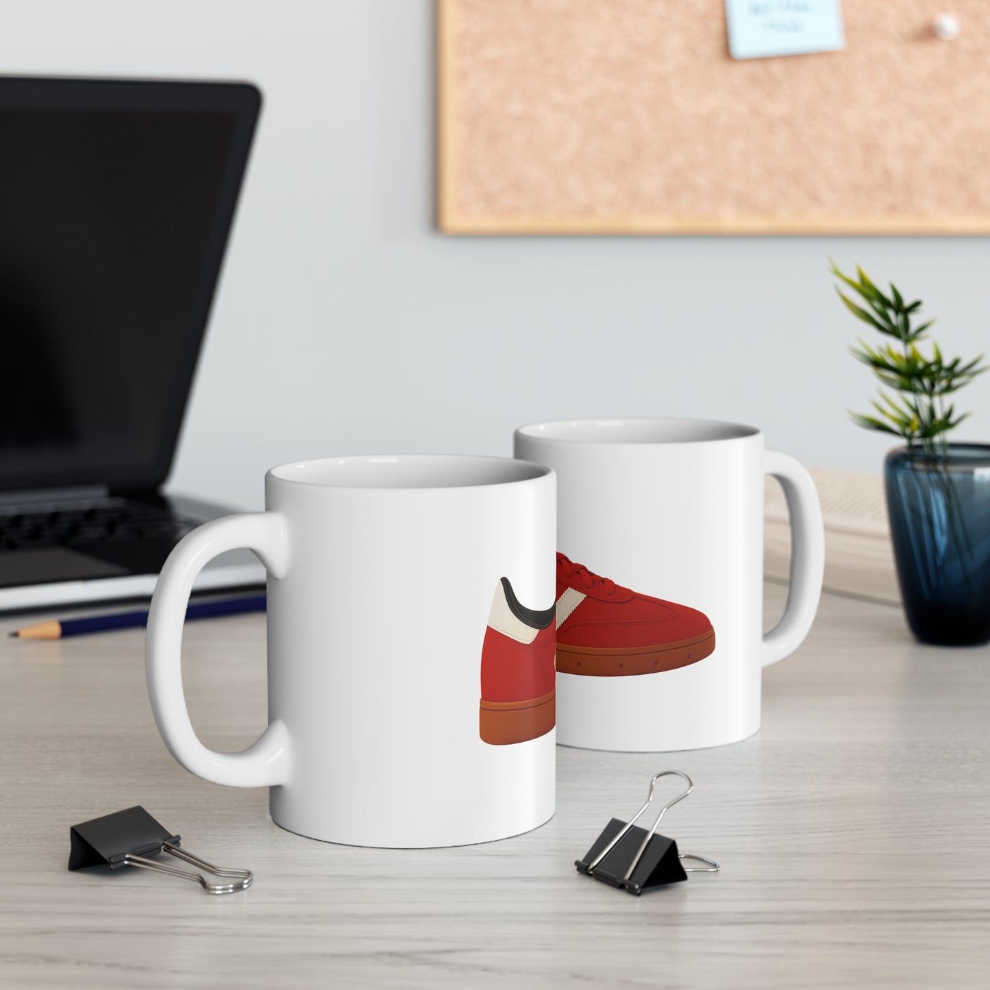 Two white ceramic mugs on a desk, each displaying a red and white sneaker design, with office supplies and laptop nearby