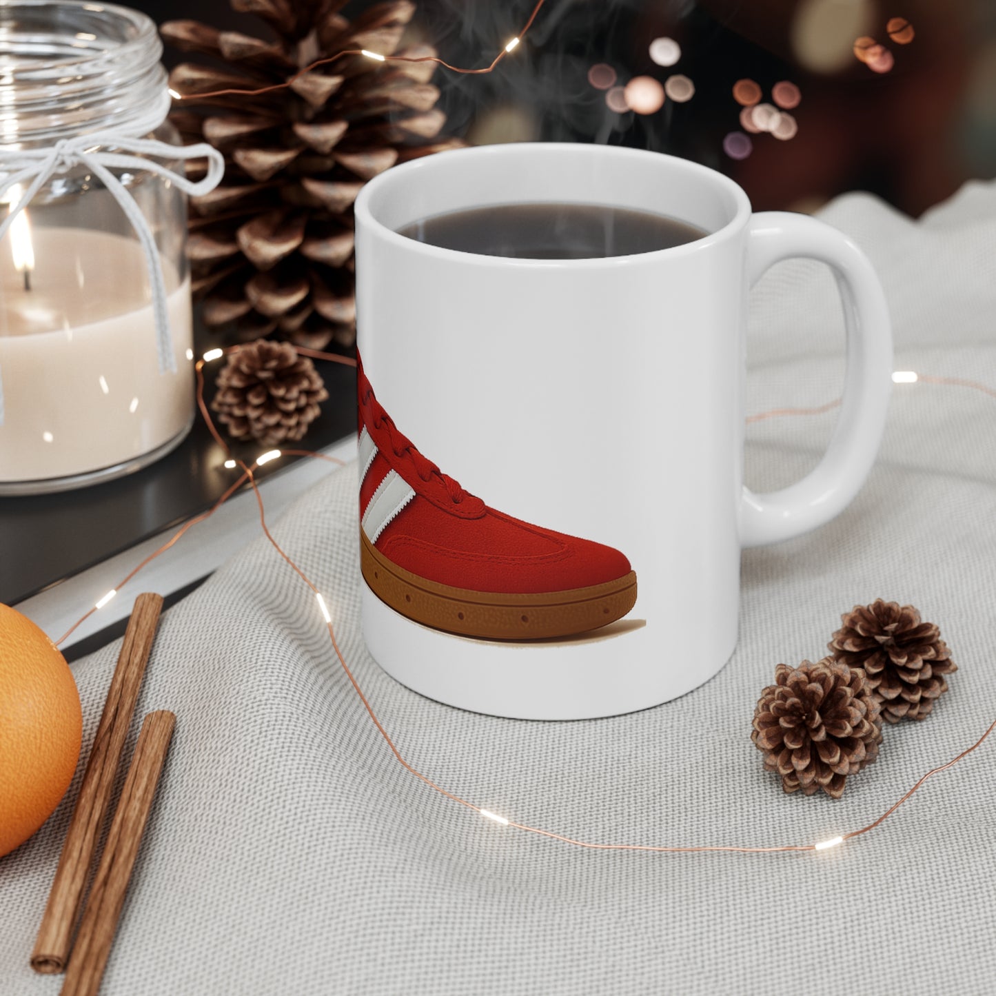 White mug with red sneaker print filled with coffee, surrounded by pinecones, candle, cinnamon sticks, and orange