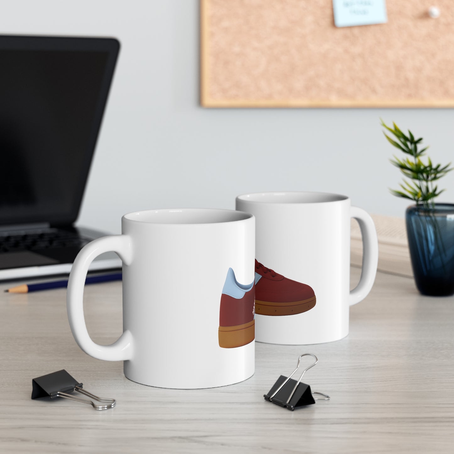 White ceramic mugs with red sneaker print on wooden desk with laptop and plant