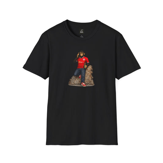 A black Barnsley short-sleeved t-shirt featuring a central graphic of a chimpanzee wearing a red football jersey with white trim, denim jeans, and red trainers. He is leaning against a section of a brick wall while holding a green bottle and talking on a mobile phone.