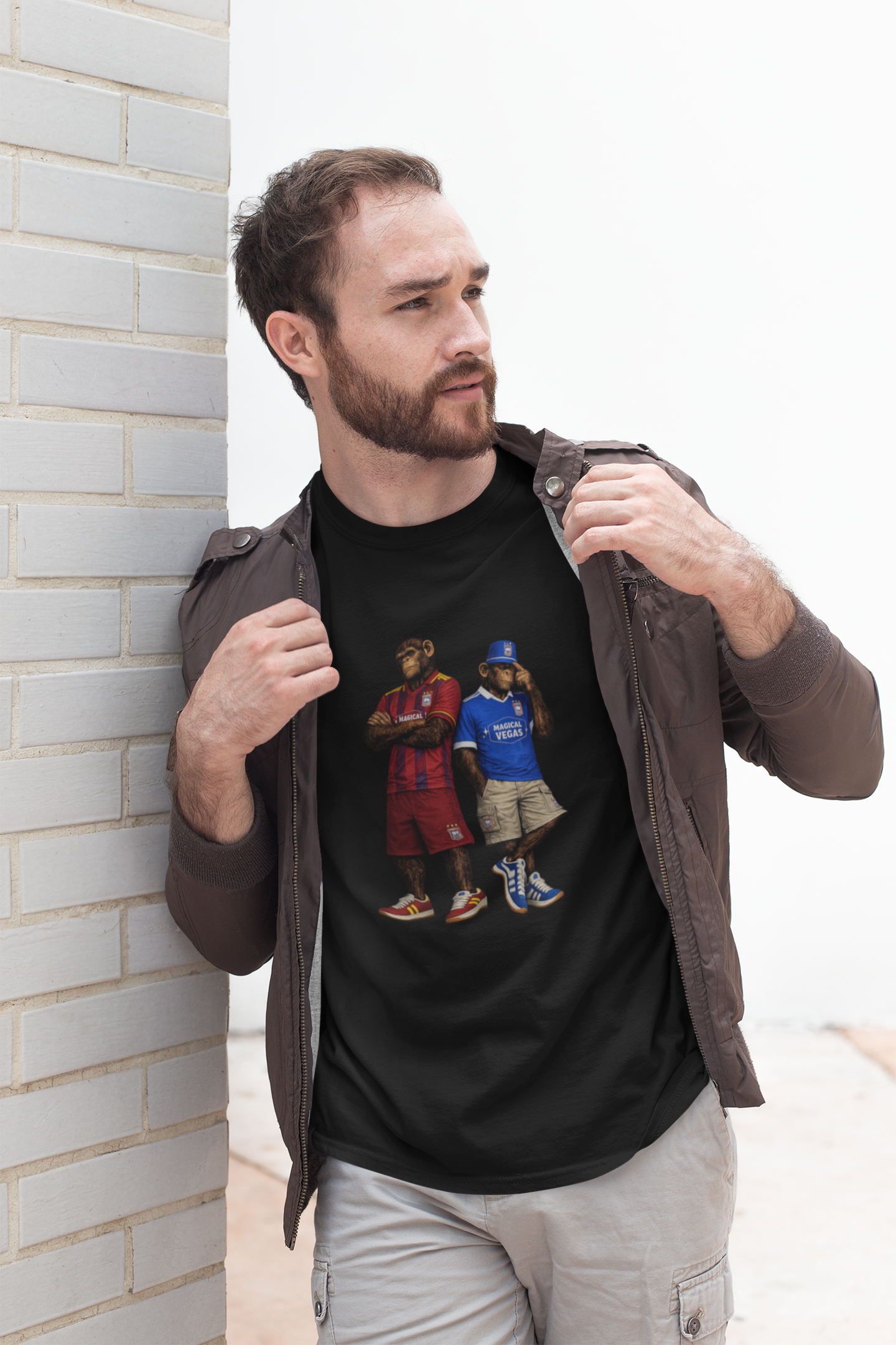 A man wearing a black Ipswich t-shirt with a detailed print of two basketball players, styled with an open brown jacket and light gray cargo pants, leaning against a white brick wall.