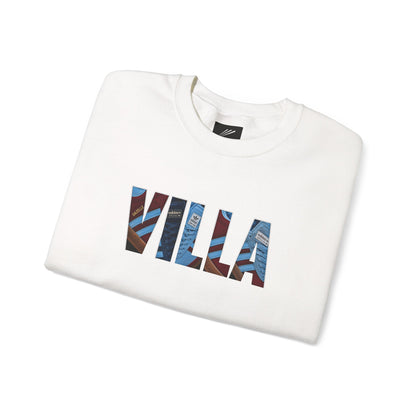 A folded white crewneck sweatshirt featuring a graphic print of the word 'VILLA' in large block letters filled with a collage of blue, red, and brown striped fashion magazine covers, set against a plain white background.