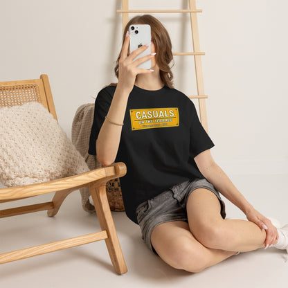 A woman sitting on the floor in a minimalist room, wearing a black Watford t-shirt with a yellow graphic box that reads 'CASUALS OUT OF OFFICE' and gray denim shorts. She is holding a white smartphone to take a mirror selfie, with a wooden chair and ladder in the background.