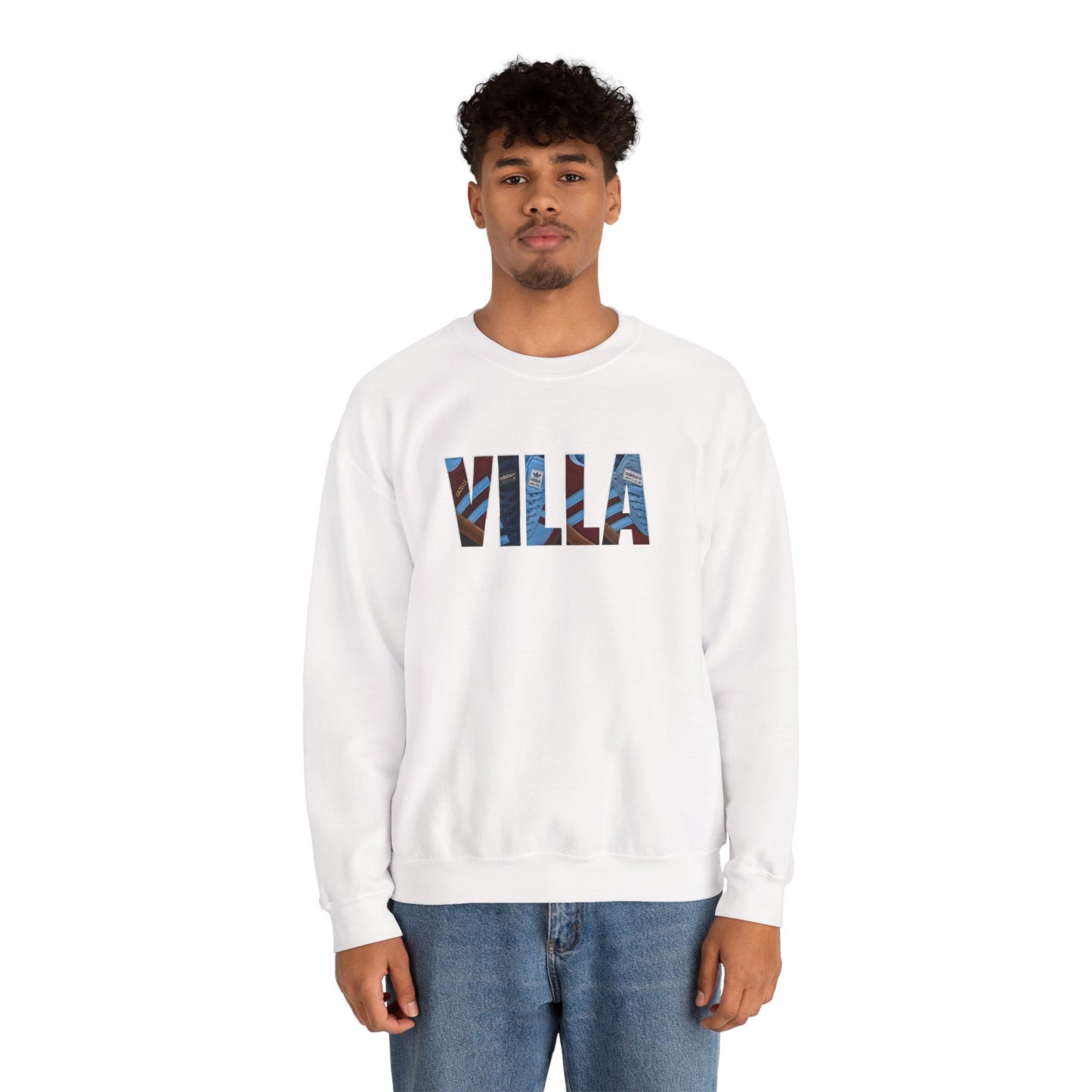 A man wearing a white crewneck sweatshirt with the word 'VILLA' printed across the chest in a blue and red patterned design, paired with blue jeans, against a plain white background.