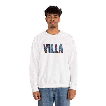 A man wearing a white crewneck sweatshirt with the word 'VILLA' printed across the chest in a blue and red patterned design, paired with blue jeans, against a plain white background.