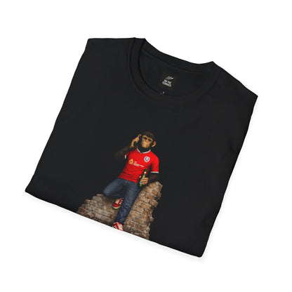 A black short-sleeved t-shirt featuring a central graphic of a chimpanzee wearing a red football jersey with white trim, denim jeans, and red trainers. He is leaning against a section of a brick wall while holding a green bottle and talking on a mobile phone.
