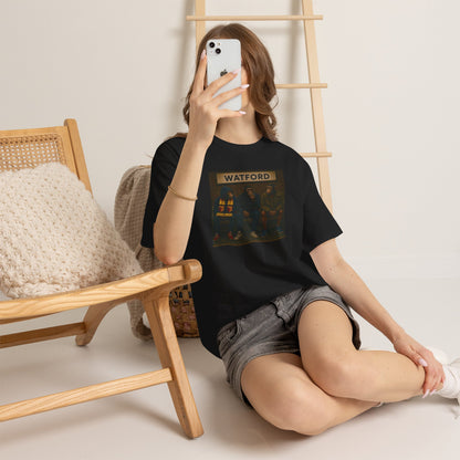 Person wearing a black Watford t-shirt with a graphic design, sitting on the floor with a phone in front of their face.