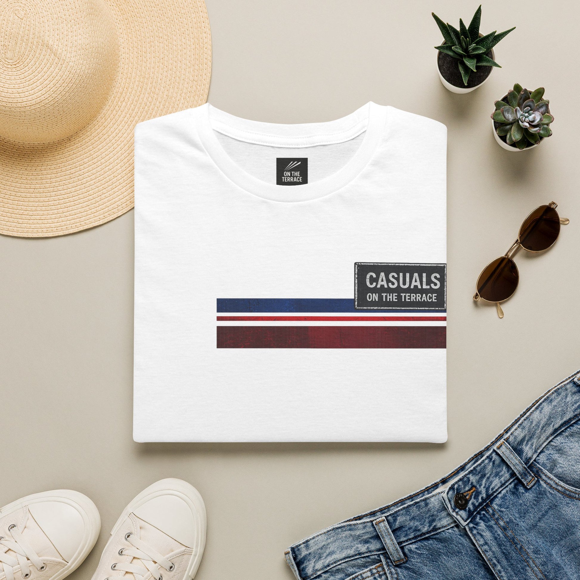 White t-shirt with 'Casuals on the Terrace' design, surrounded by a hat, sunglasses, shoes, and plants on a beige surface.
