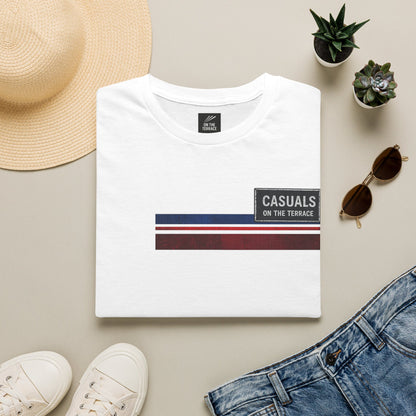 White t-shirt with 'Casuals on the Terrace' design, surrounded by a hat, sunglasses, shoes, and plants on a beige surface.