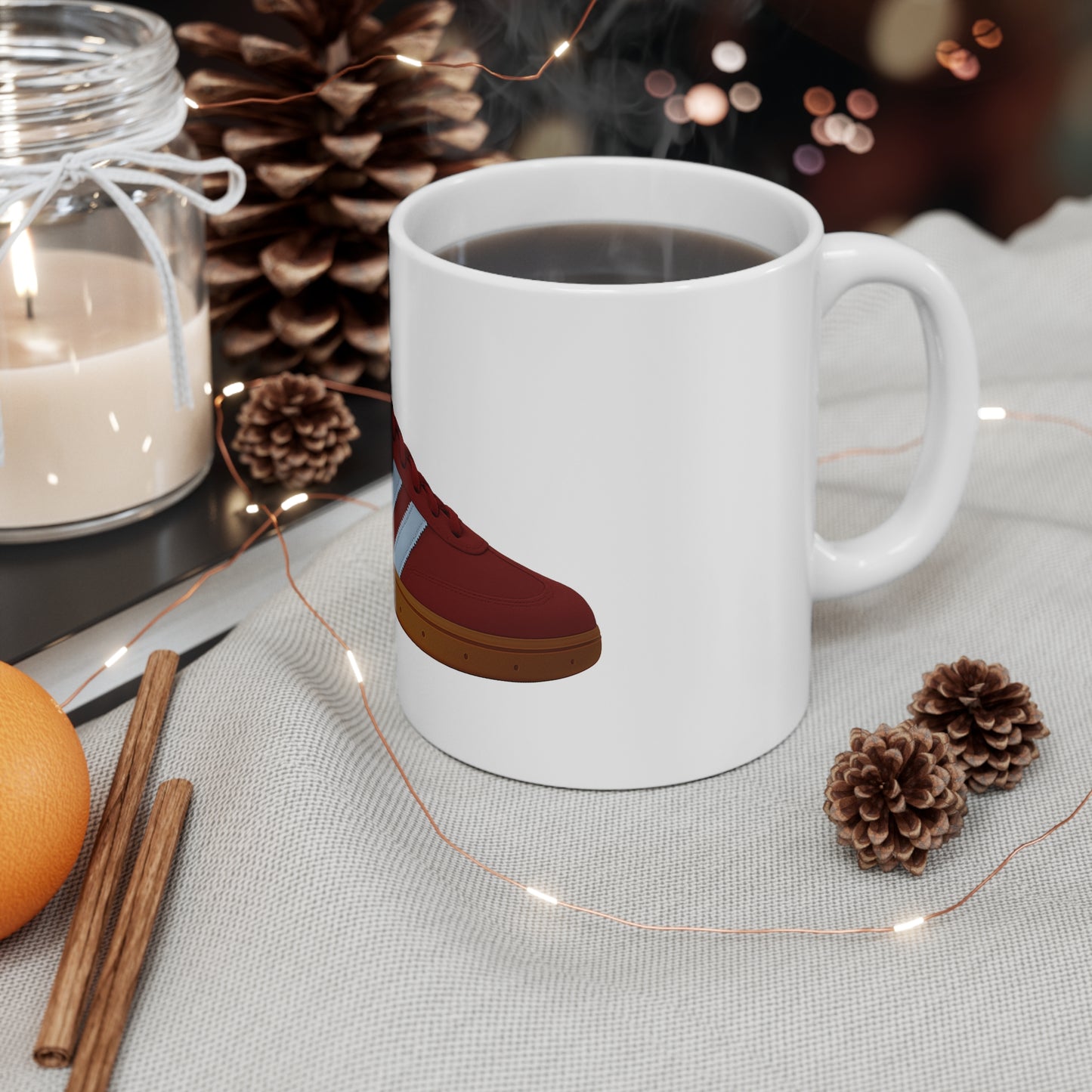 White coffee mug with red sneaker graphic on cozy fabric beside pine cones, cinnamon sticks, orange, candle, and fairy lights