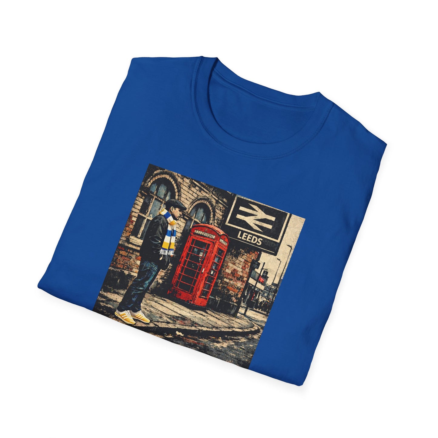 A royal blue T-shirt folded neatly against a white background. On the front is a square printed image showing a man standing on a worn city pavement. He wears a dark jacket, flat cap, jeans, a blue-and-yellow striped scarf, and bright yellow trainers. Behind him is an old brick building with arched windows, a red British telephone box, and a black-and-white railway sign that reads “LEEDS.” The overall scene has a gritty, urban, vintage feel.