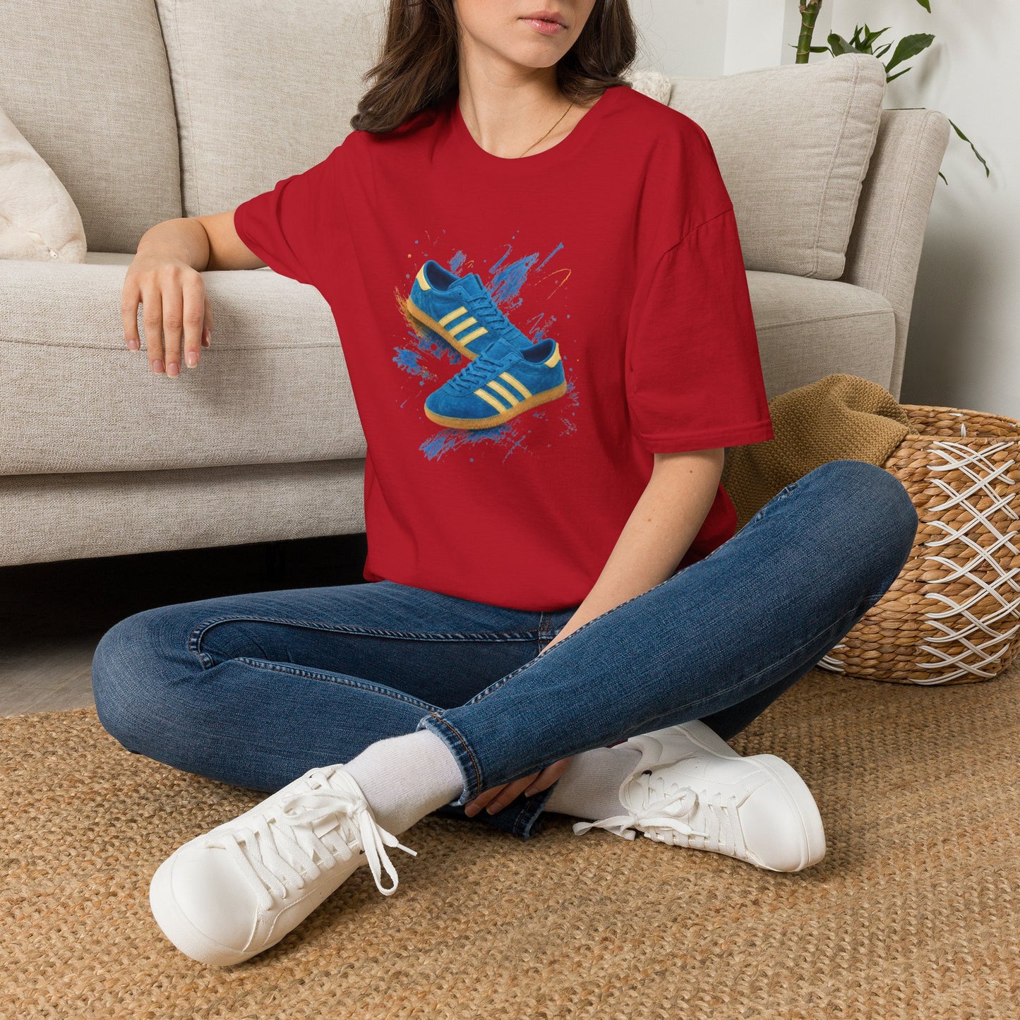 A woman sitting cross-legged on a woven rug in front of a beige sofa, wearing a red graphic t-shirt with a blue and yellow sneaker design, blue jeans, and white sneakers.