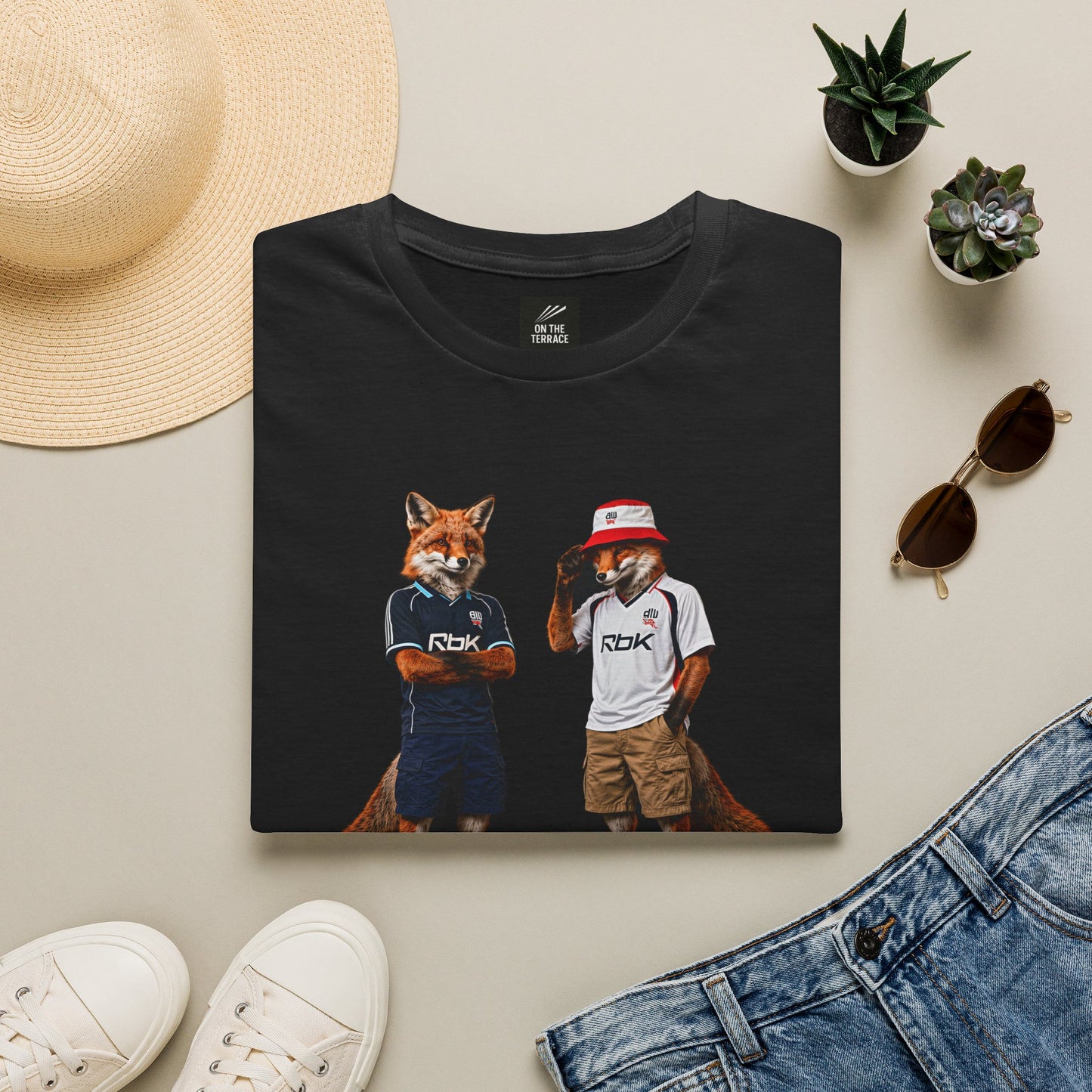 A black graphic t-shirt laid flat, featuring a central design by On The Terrace for Bolton fans. The graphic showcases two anthropomorphic English foxes dressed in vintage Rbk Bolton-inspired football kits. The fox on the left wears the dark blue retro away jersey with arms crossed, while the fox on the right sports the classic white home shirt, a red and white bucket hat, and cargo shorts. The inner neck of the tee features the official "On The Terrace" brand logo