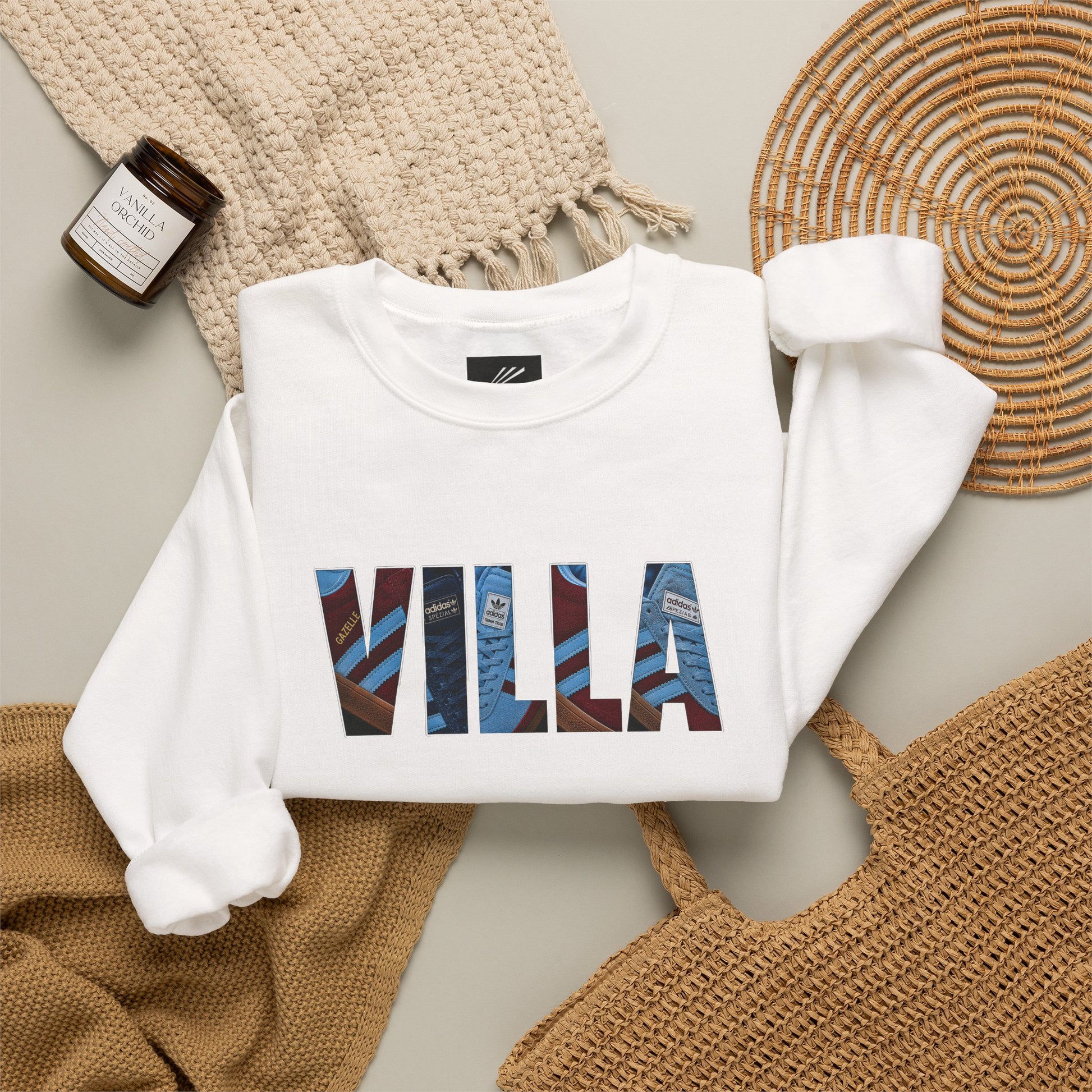 A folded white sweatshirt with the word 'VILLA' printed across the chest in large blue and burgundy patterned letters, styled in a flat lay with a woven blanket, a small candle jar, a wicker tray, and a tan tote bag on a light neutral background.