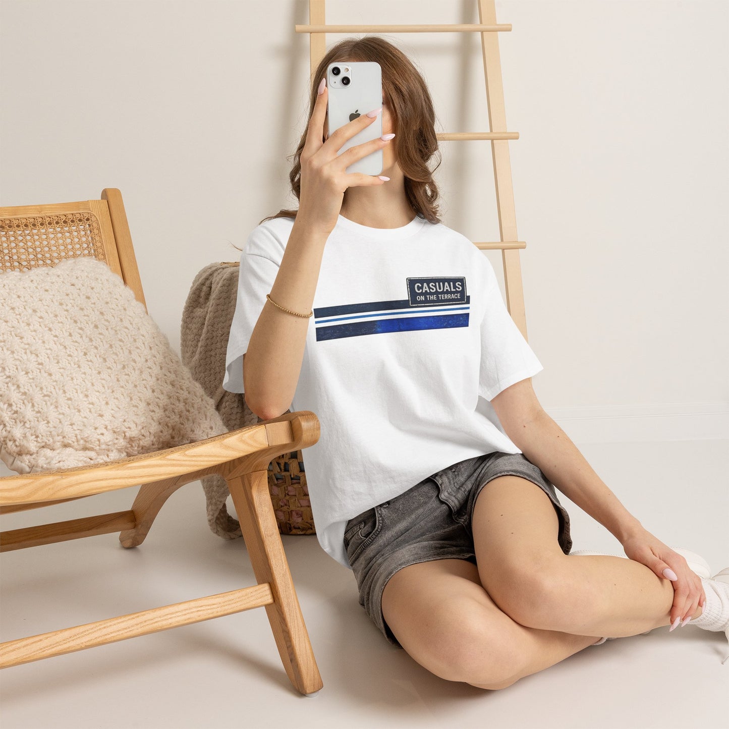 Person wearing a white t-shirt with a graphic design, sitting on the floor with a wooden chair and ladder in the background.