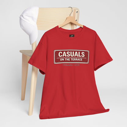 A red heavy cotton Aberdeen t-shirt flat lay featuring a central vintage red and white street sign graphic reading "CASUALS ON THE TERRACE" with "Pittodrie • 1903" printed below, and an inner neck label for On The Terrace. hanging on a wooden hanger from a light wood chair. A white baseball cap rests on the back of the chair against a clean, light gray studio background.
