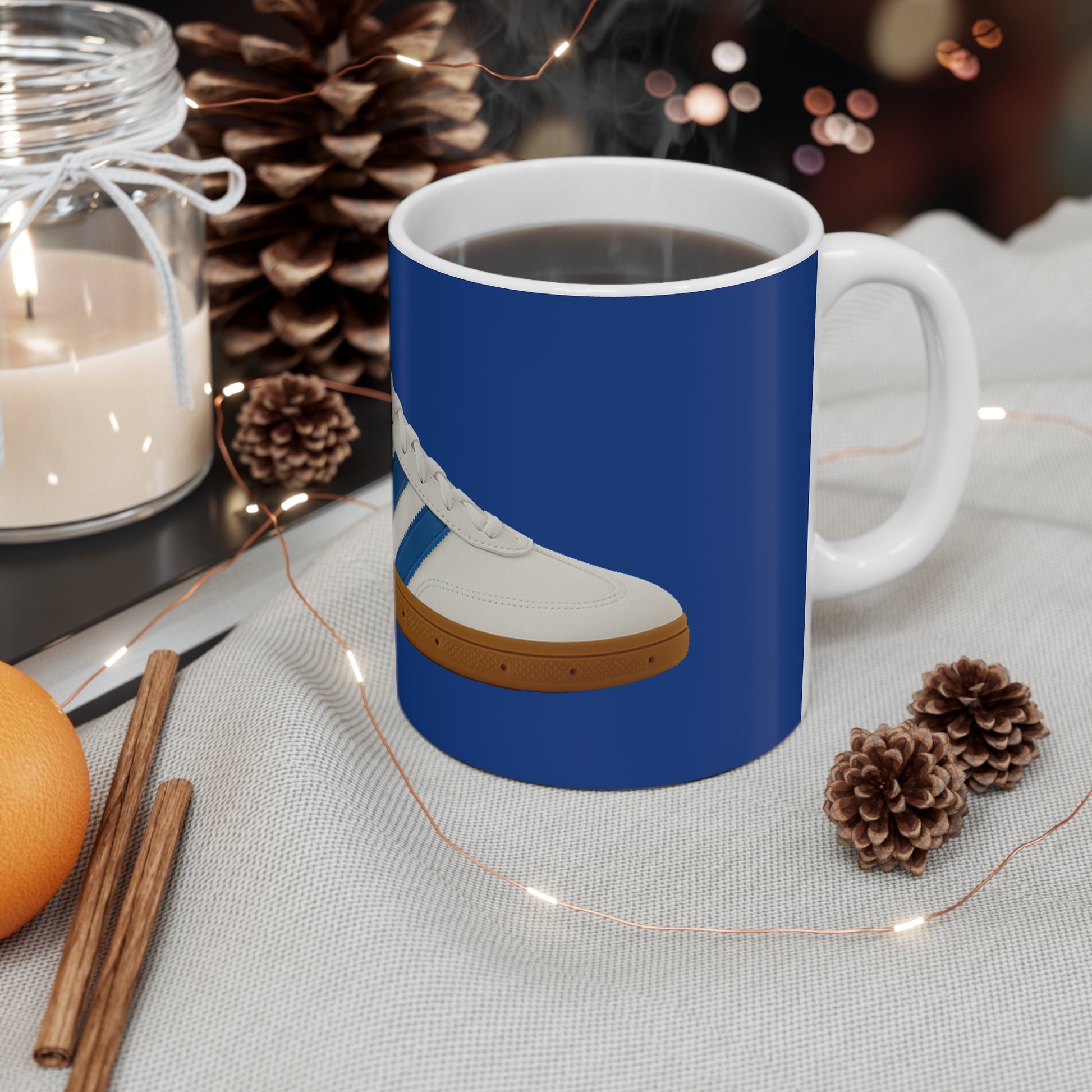 Steaming coffee mug with blue sneaker design, surrounded by pinecones, cinnamon sticks, orange, candle, and string lights on fabric