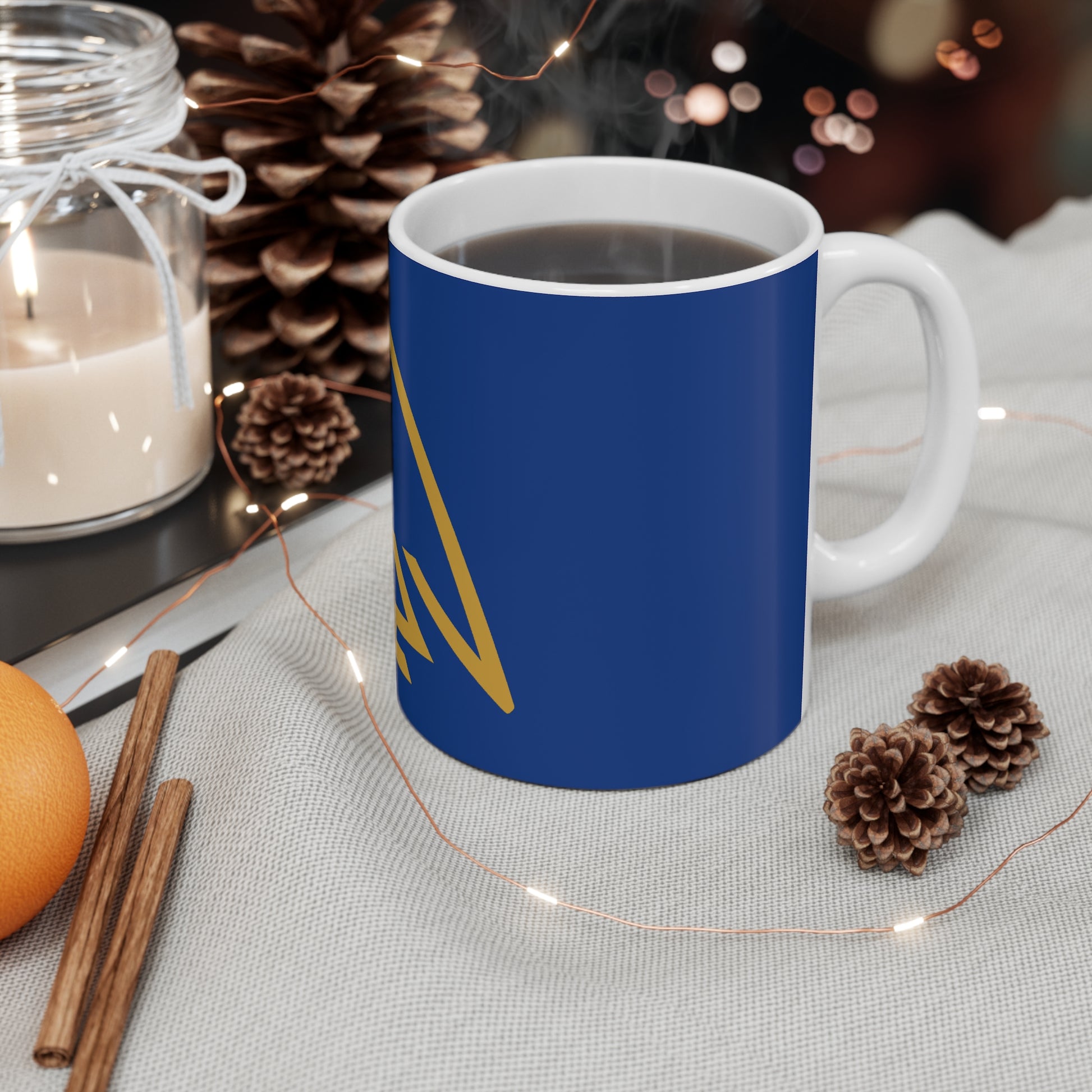 Blue coffee mug with gold design steaming hot coffee on textured cloth with pine cones, cinnamon sticks, candle
