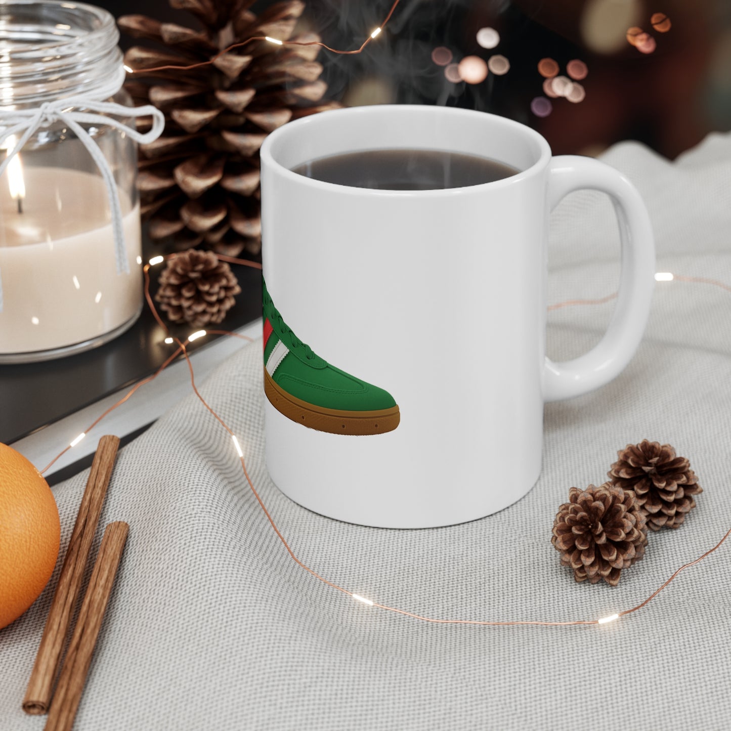 Steaming black coffee in white mug with green sneaker graphic, surrounded by pinecones, cinnamon sticks, candle