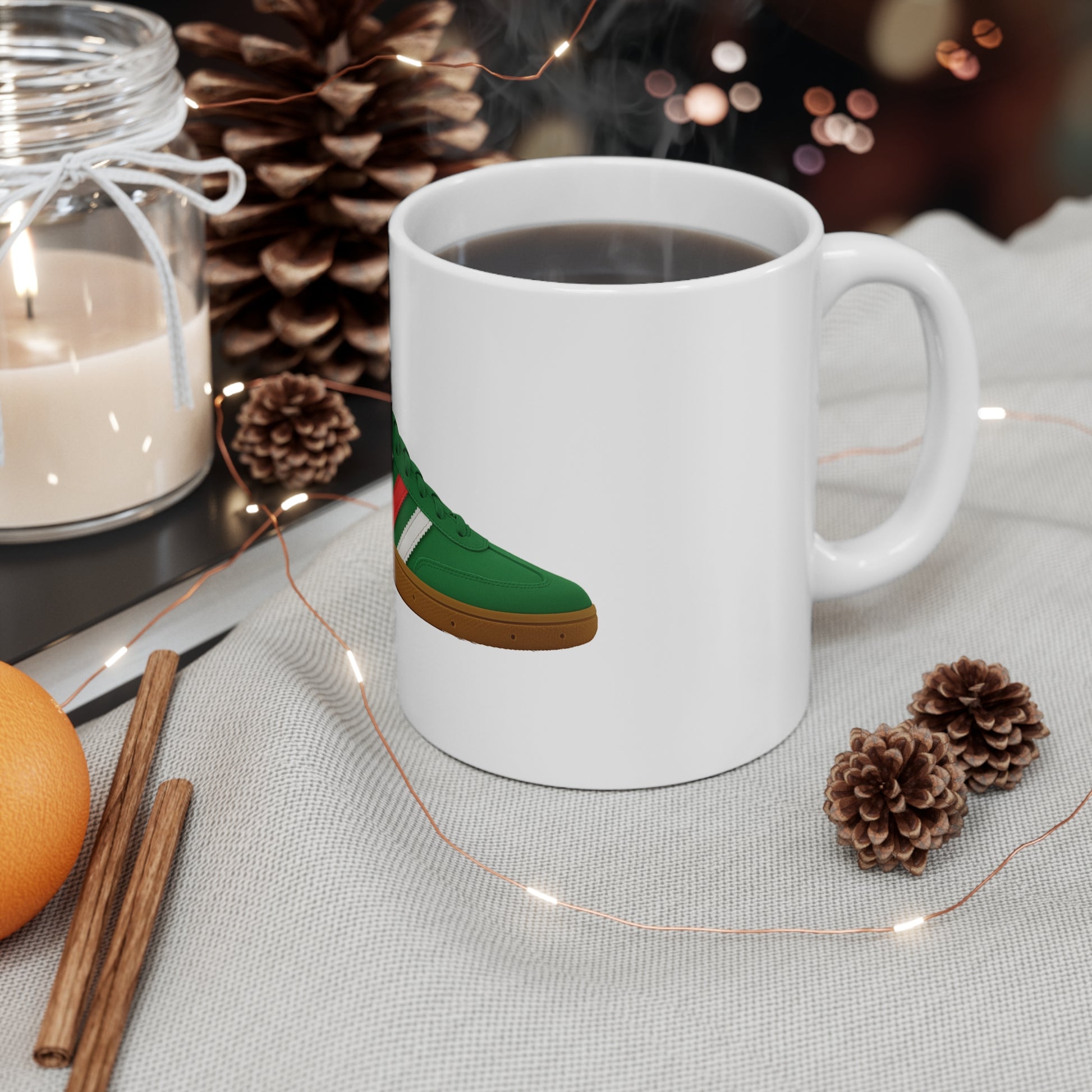 Steaming black coffee in white mug with green sneaker graphic, surrounded by pinecones, cinnamon sticks, candle