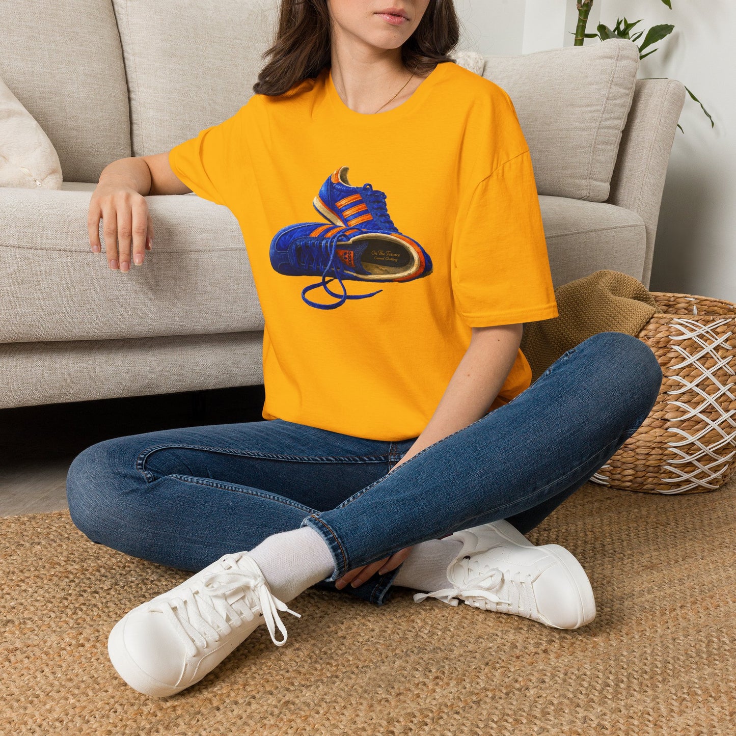 A woman sitting cross-legged on a woven rug in front of a light beige sofa, wearing an Gold t-shirt with hand-illustrated blue and orange retro terrace trainer sneaker graphic print, On The Terrace casual clothing brand
