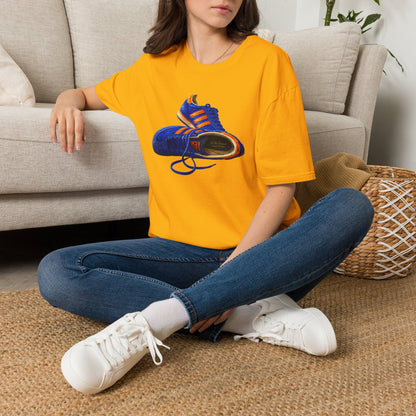 A woman sitting cross-legged on a woven rug in front of a light beige sofa, wearing an Gold t-shirt with hand-illustrated blue and orange retro terrace trainer sneaker graphic print, On The Terrace casual clothing brand

