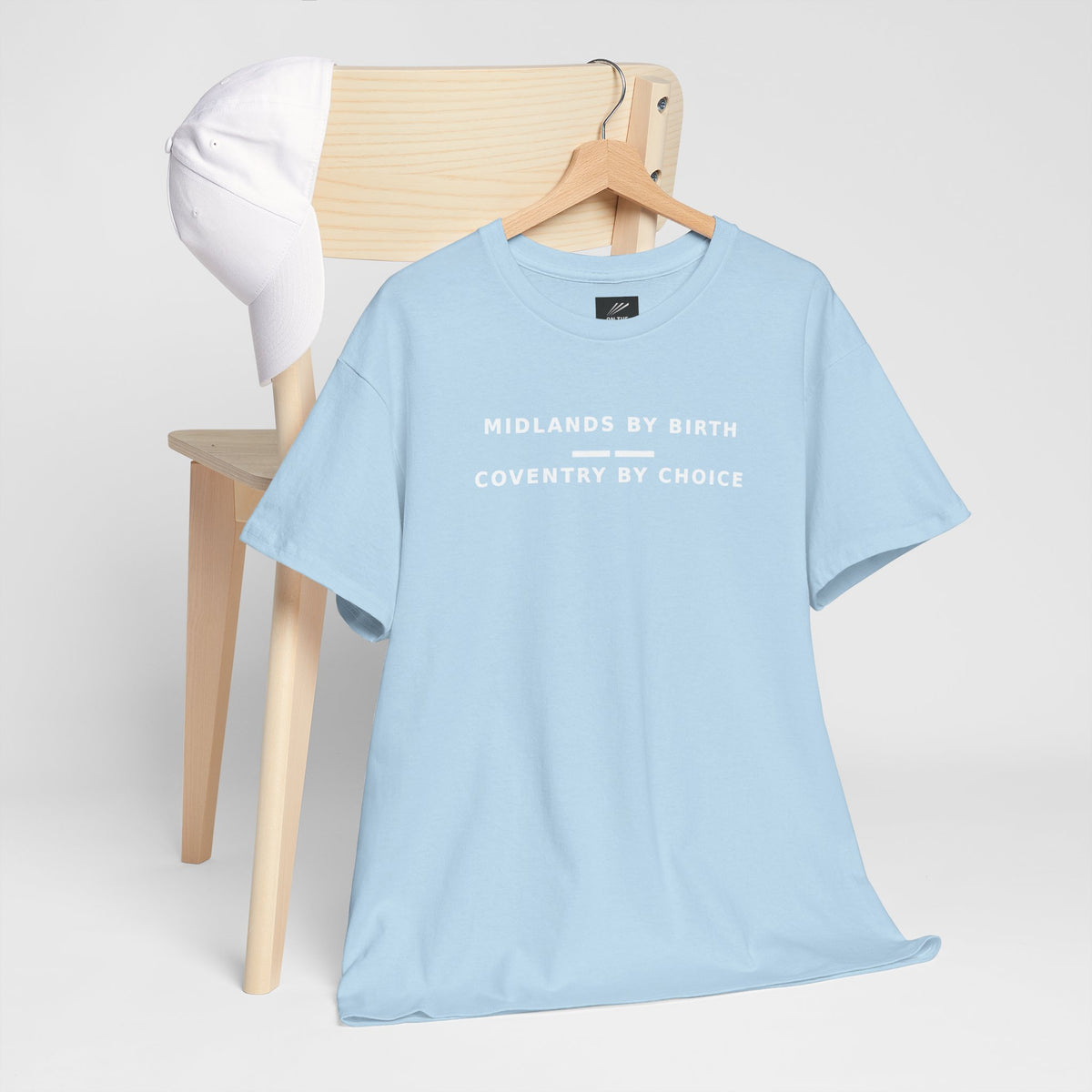 Sky blue Midlands By Birth, Coventry By Choice T-shirt hanging by a wooden chair