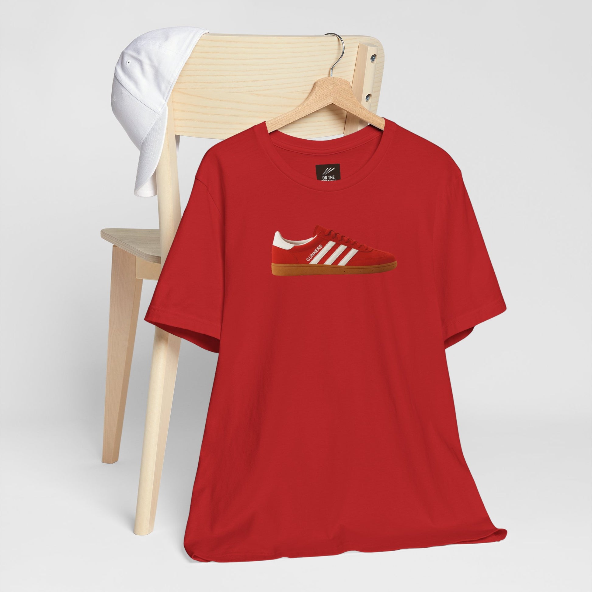 Red t-shirt with red and white sneaker graphic hanging on wooden chair with white cap