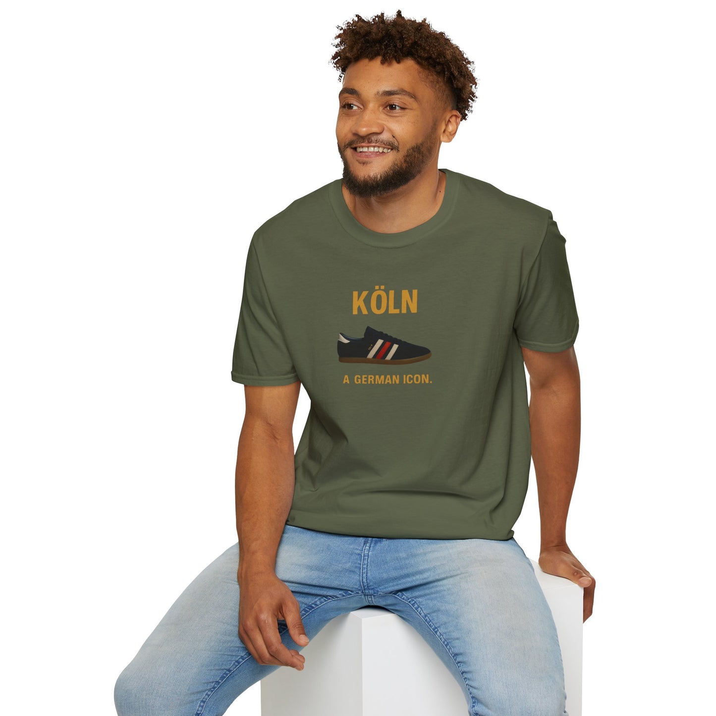 Smiling man wearing olive green t-shirt with Köln shoe print and text 'A German icon.' sitting on white cube