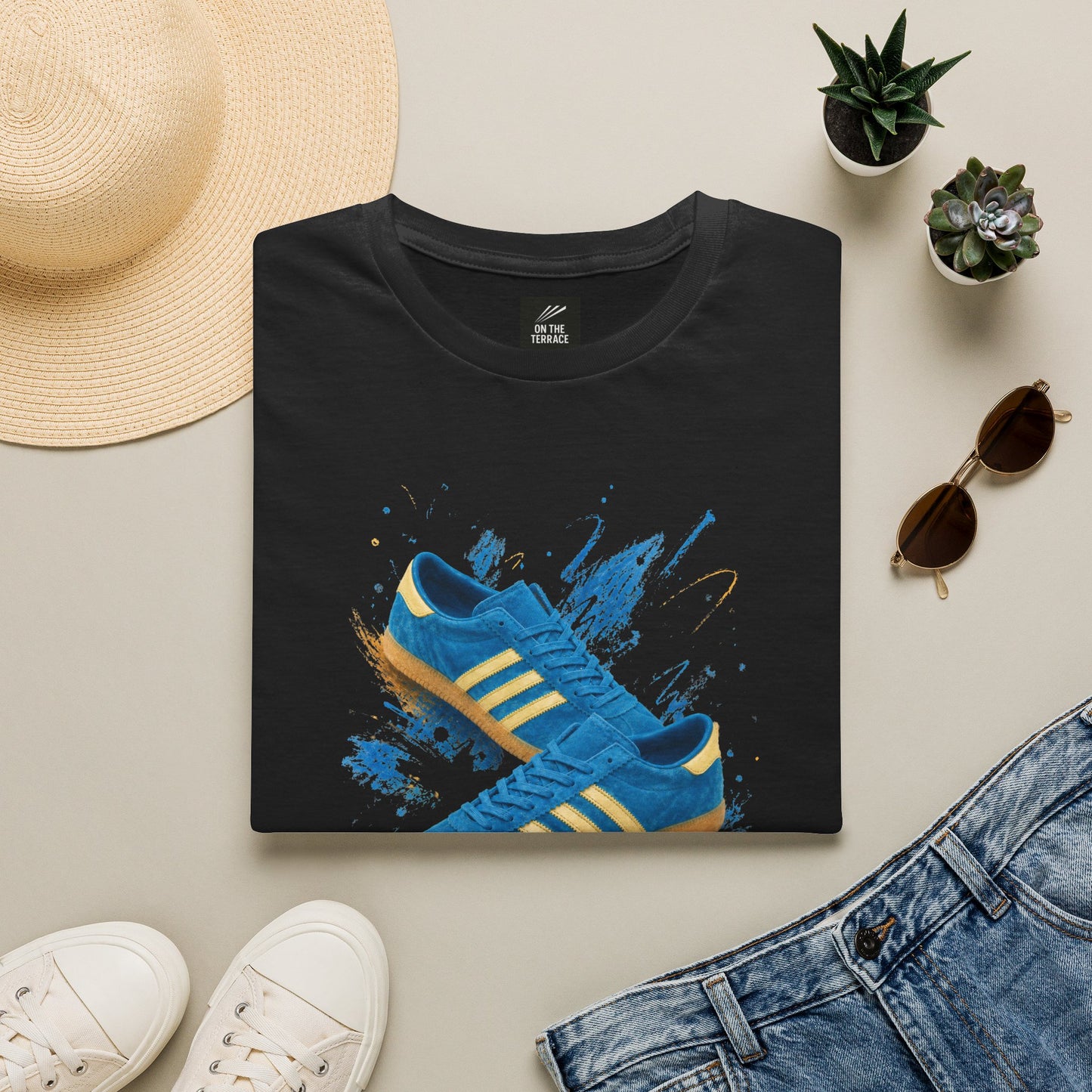 A folded black T-shirt is displayed in a styled flat lay on a light beige surface, featuring a graphic of blue sneakers with yellow stripes on a blue paint-splatter background. Surrounding props include a straw hat, white sneakers, blue jeans, small potted succulents, and sunglasses.
