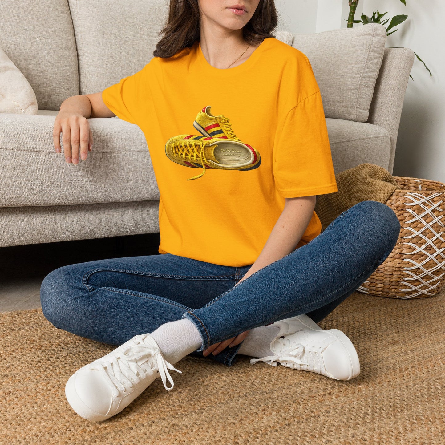 A woman sitting cross-legged on a woven rug in front of a beige sofa, wearing an Gold t-shirt with hand-illustrated yellow, navy and red retro terrace trainer sneaker graphic print, On The Terrace casual clothing brand

