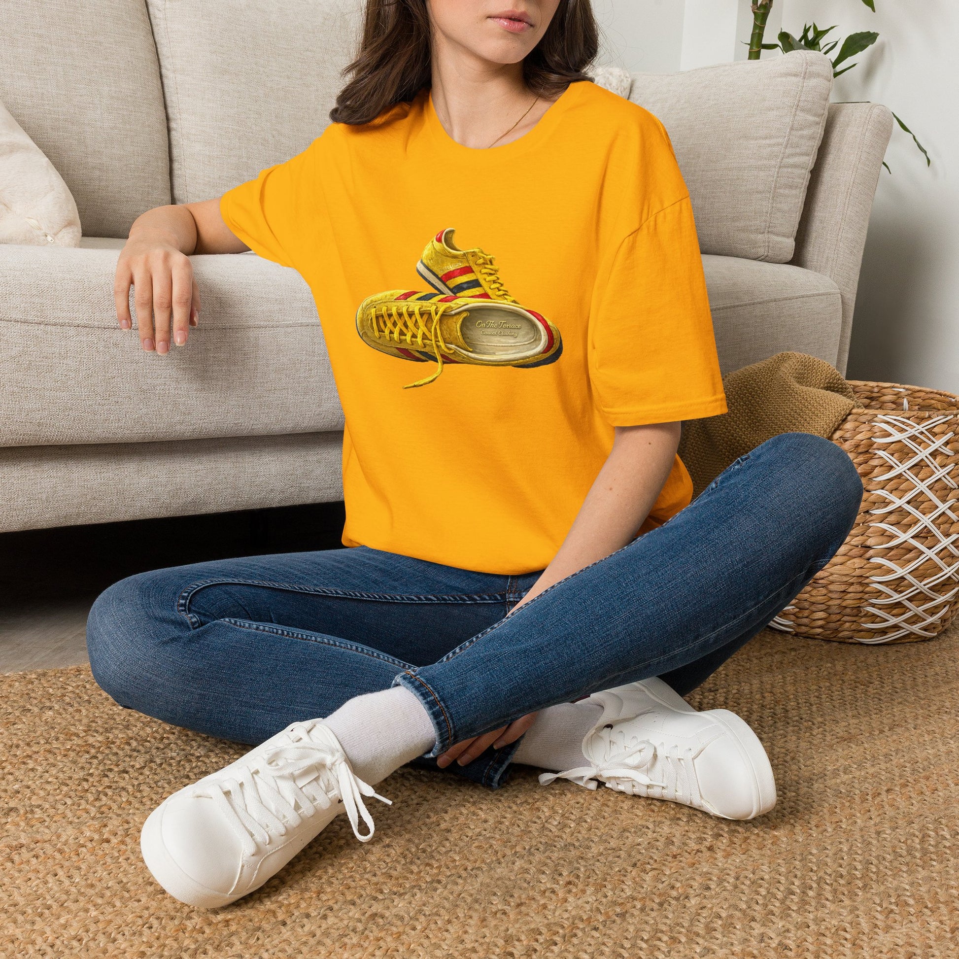 A woman sitting cross-legged on a woven rug in front of a beige sofa, wearing an Gold t-shirt with hand-illustrated yellow, navy and red retro terrace trainer sneaker graphic print, On The Terrace casual clothing brand
