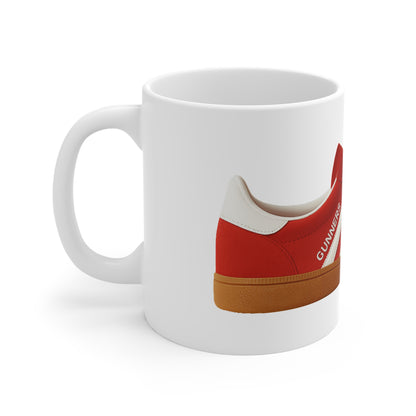 A white ceramic coffee mug featuring a graphic print from the file from On The Terrace, showing a classic red retro terrace trainer with three white stripes, a brown gum sole, and the word "GUNNERS" printed on the side.