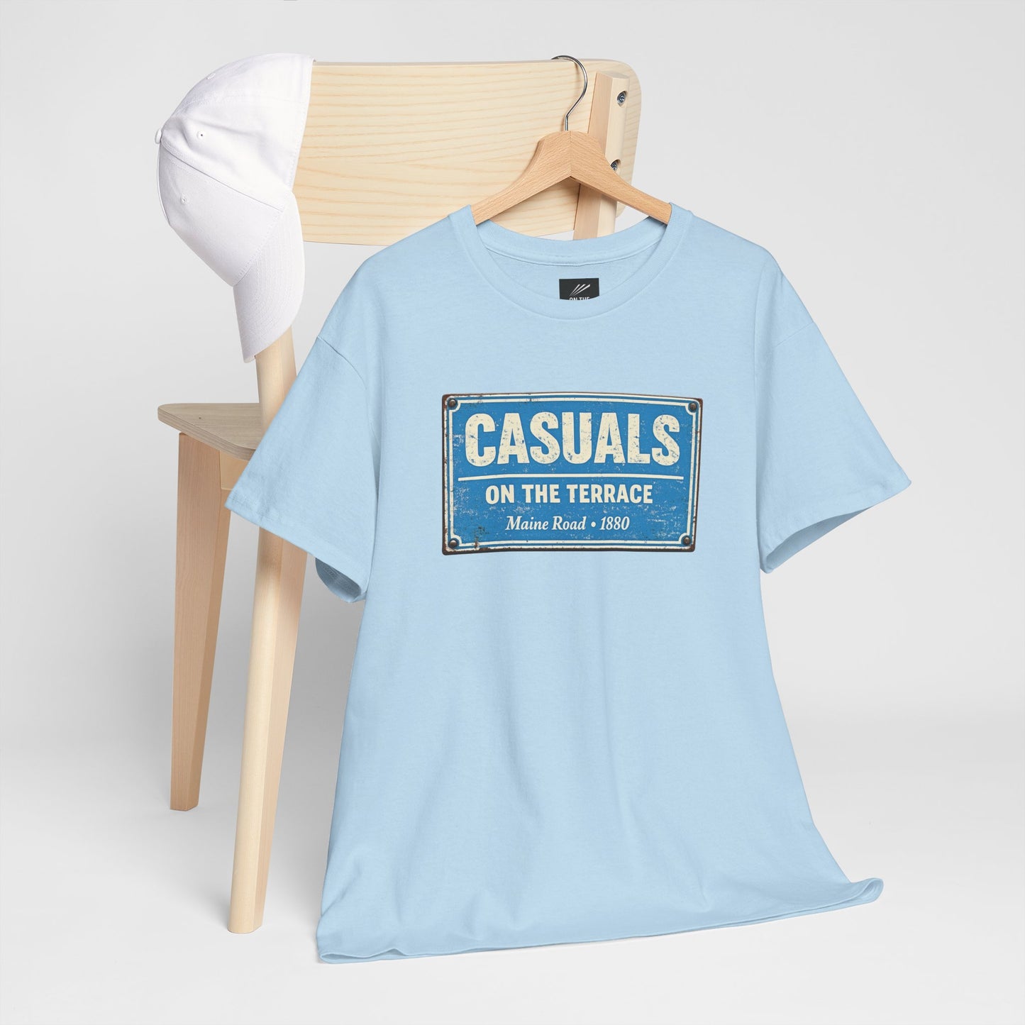 A light blue MCFC t-shirt hanging on a wooden hanger from a light wood chair, featuring a distressed blue and white vintage-style sign graphic on the chest that reads 'CASUALS ON THE TERRACE'.