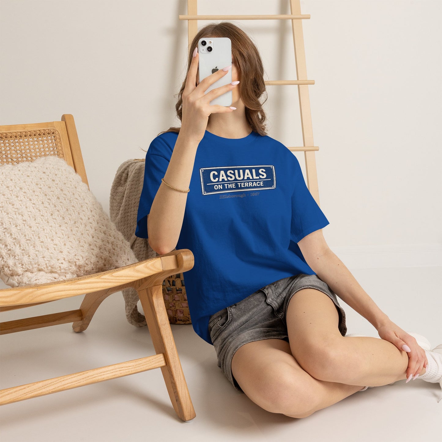 Person wearing a royal blue heavy cotton  SWFC t-shirt flat lay featuring a central vintage blue and white street sign graphic reading "CASUALS ON THE TERRACE" with "Hillsborough • 1867" printed below, and an inner neck label for On The Terrace, sitting on the floor with a phone