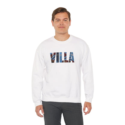 A man wearing a white crewneck sweatshirt with the word 'VILLA' printed across the chest in a blue and brown scenic pattern, paired with dark gray pants, against a plain white background.