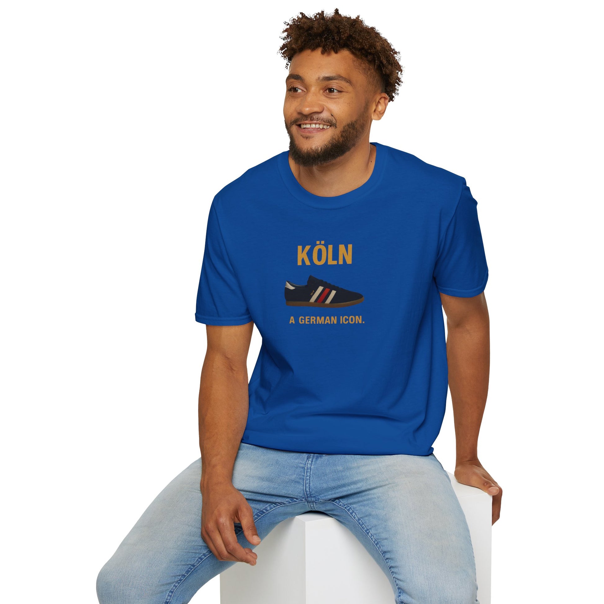 Smiling man wearing a blue t-shirt with Köln German icon sneaker design, sitting on white cube