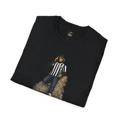 A folded black short-sleeved crew-neck t-shirt featuring a central graphic of a chimpanzee dressed in a black and white striped football jersey, denim jeans, and black trainers. The character is leaning against a section of a brick wall while holding a green bottle and talking on a mobile phone.