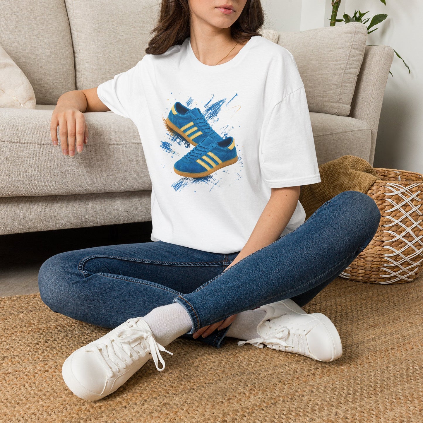 A woman sitting cross-legged on a woven rug in front of a beige sofa, wearing a white graphic T-shirt with a blue and yellow sneaker print, blue jeans, and white sneakers.