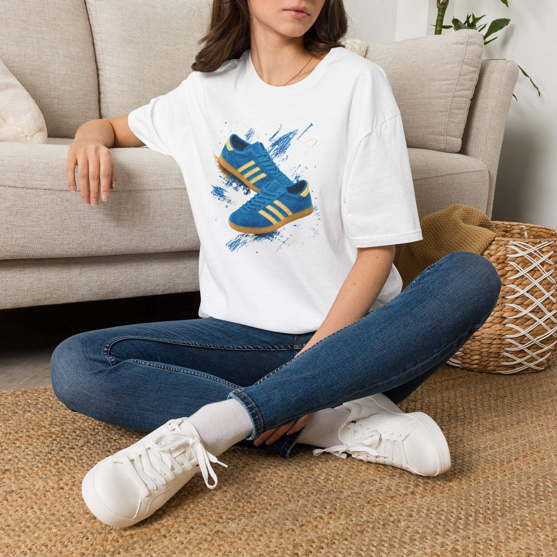 A woman sitting cross-legged on a woven rug in front of a beige sofa, wearing a white graphic T-shirt with a blue and yellow sneaker print, blue jeans, and white sneakers.