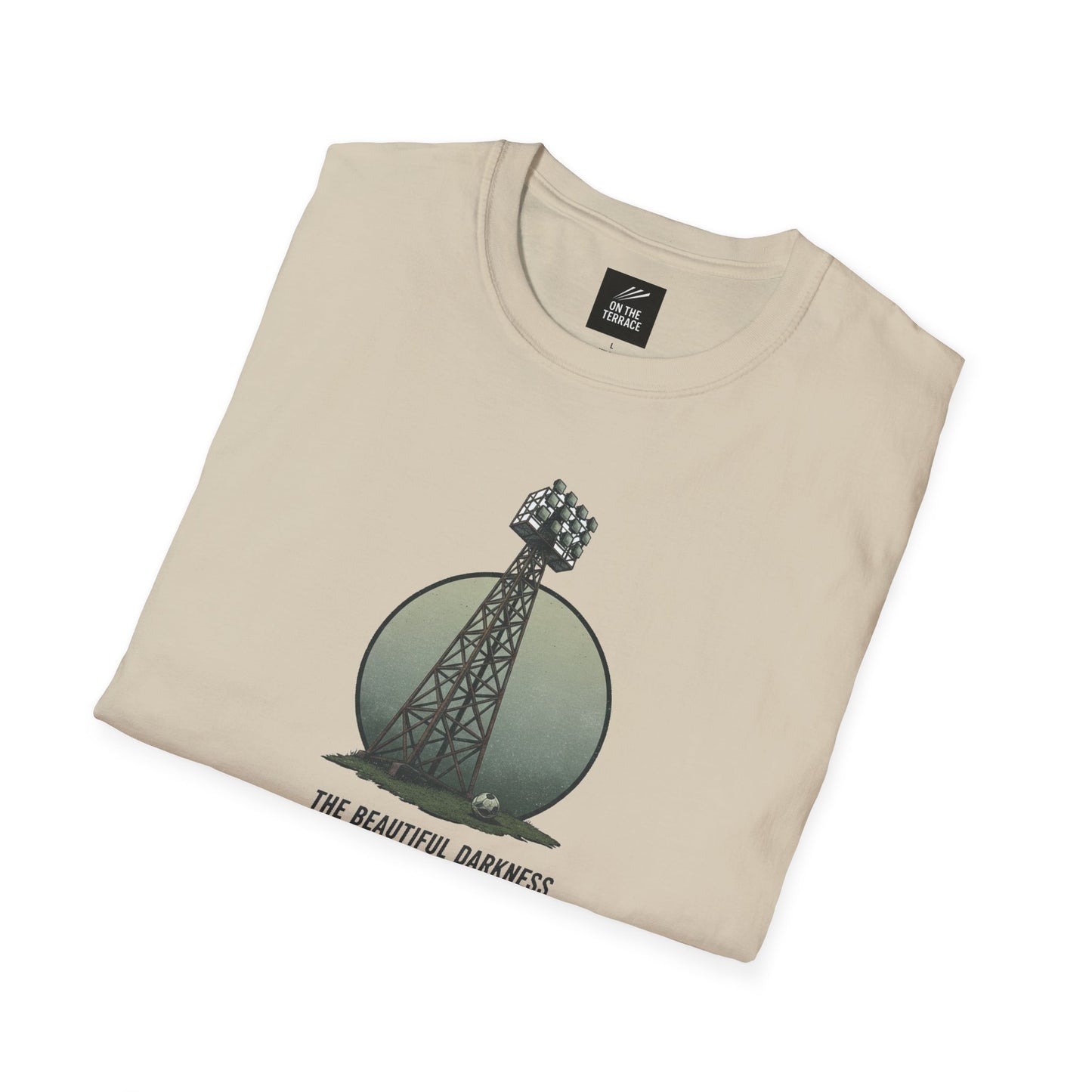 Beige folded t-shirt with a graphic of a stadium floodlight tower and soccer ball, text 'The Beautiful Darkness'