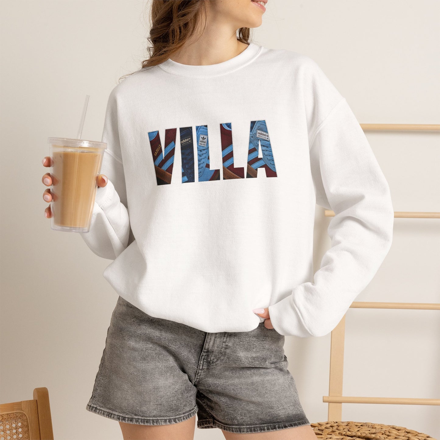 A woman wearing a white crewneck sweatshirt with a graphic text print reading 'VILLA' filled with blue and brown architectural patterns, paired with gray denim shorts and holding an iced coffee.