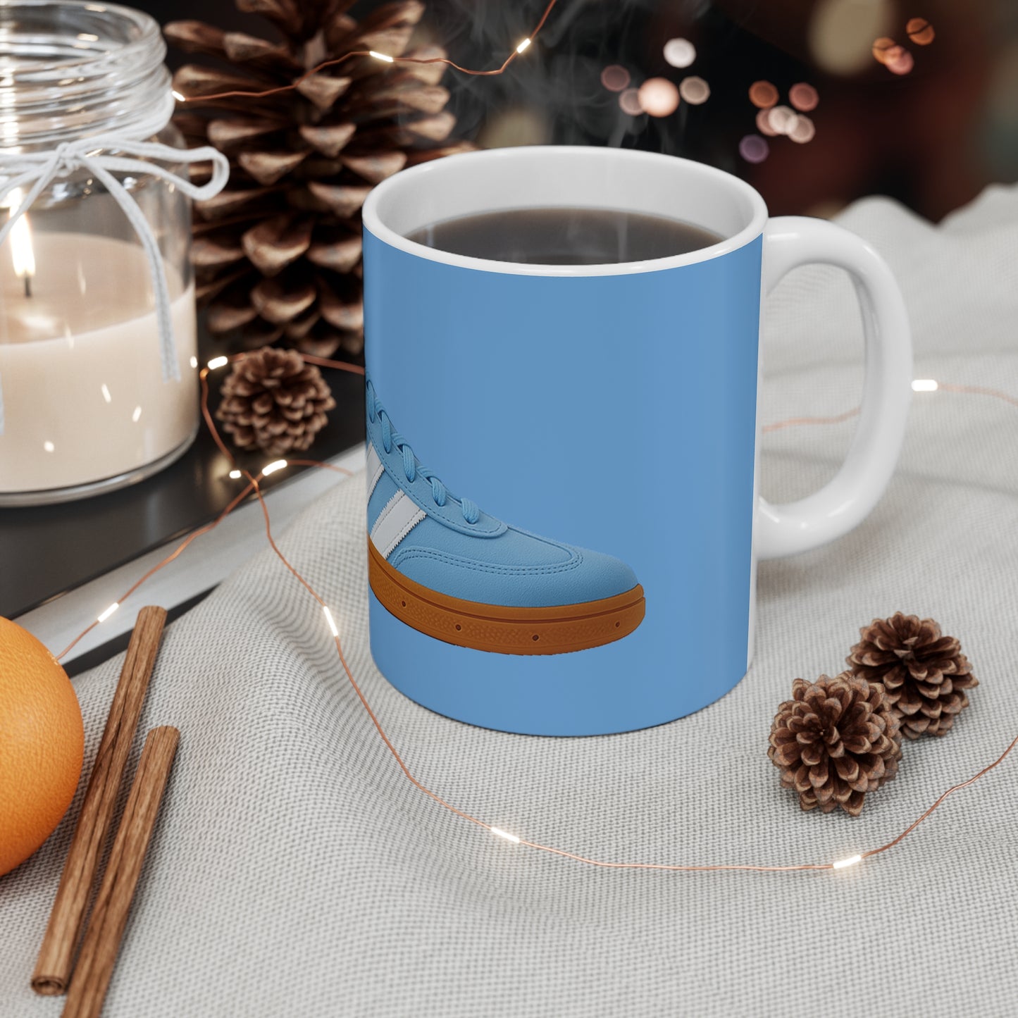 Steaming black coffee in white mug with blue sneaker design on cozy table with pine cones, cinnamon, candle, orange
