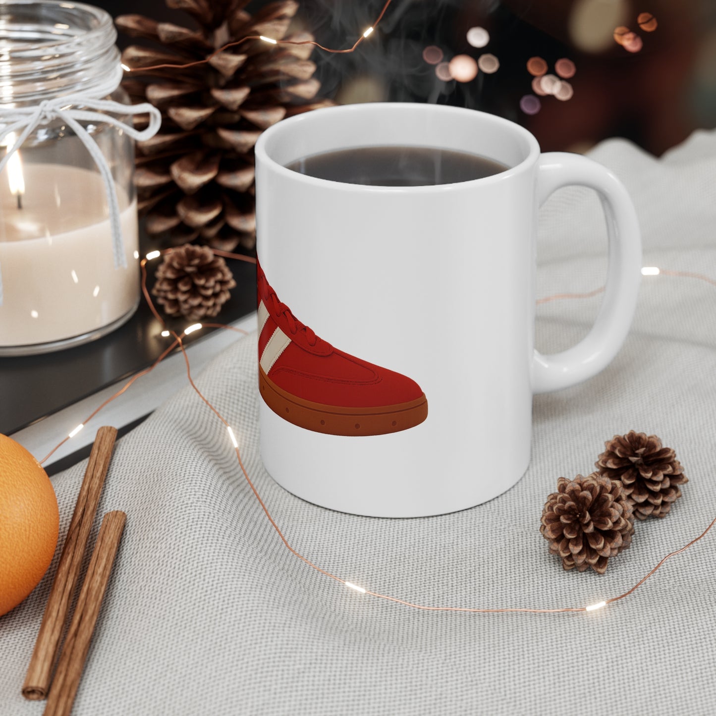 White ceramic mug with red sneaker design filled with black coffee on cozy fabric with pinecones, cinnamon sticks, and lit candle