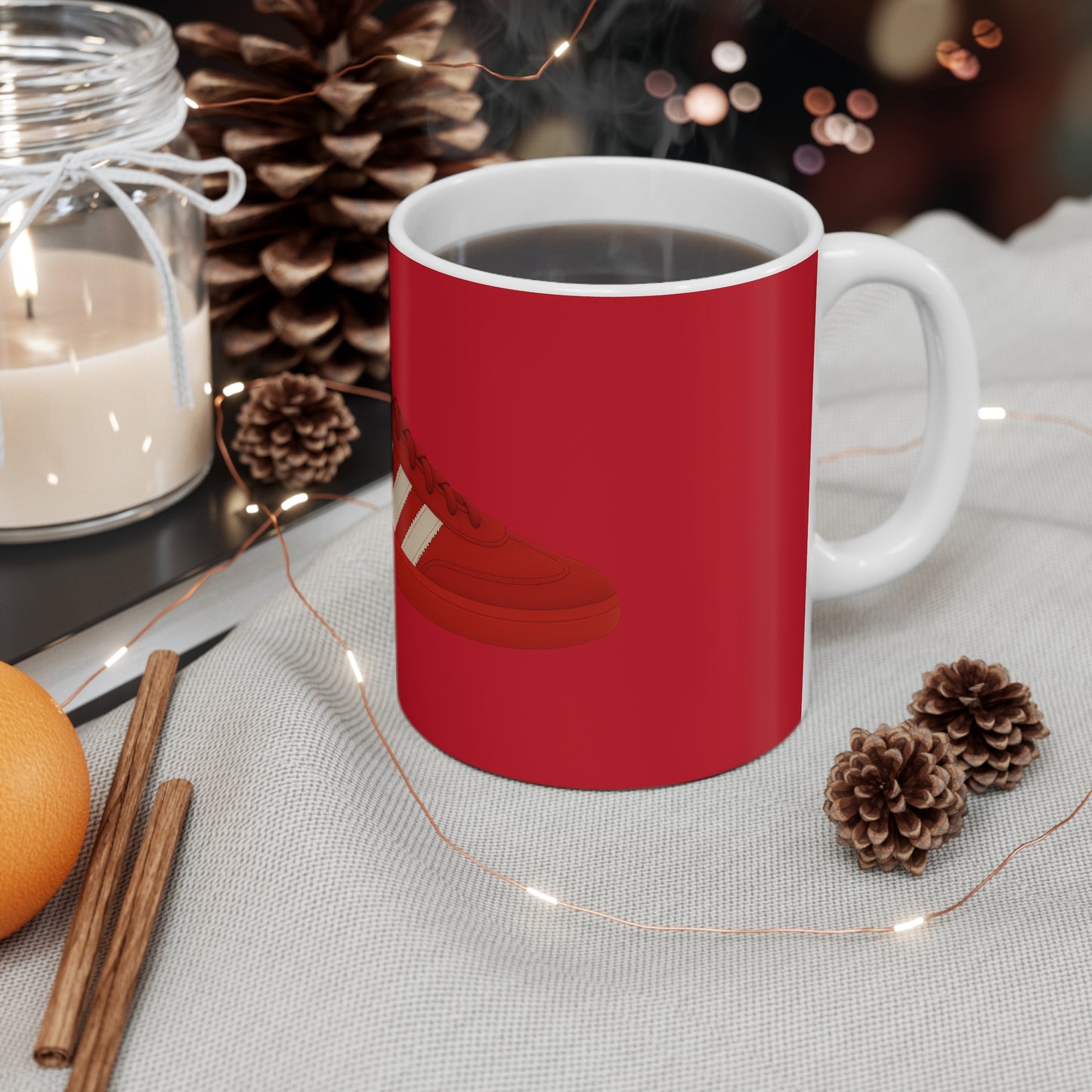Red coffee mug with sneaker design on a cozy table, surrounded by pine cones, cinnamon sticks, candle, and string lights