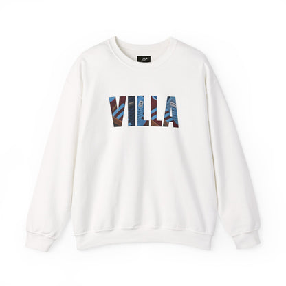 An off-white crewneck sweatshirt featuring the word 'VILLA' printed across the chest in a stylized block font filled with a blue and brown patterned graphic, set against a plain white background.